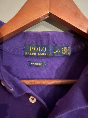 Lila pikétröja från Polo Ralph Lauren - Säljer en lila pikétröja från Polo Ralph Lauren i storlek L. Tröjan har klassisk krage, korta ärmar och knappar framtill. Tillverkad i mjuk bomull och har en custom fit-passform. Perfekt för en avslappnad men ändå stilren look.