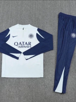 Nike Inter träningsset blå/vit - Snyggt träningsset från Nike för Inter med långärmad tröja och matchande byxor. Setet är vitt med mörkblå detaljer och har Inter-logga samt Qatar Airways-tryck på bröstet. Materialet är troligtvis polyester, perfekt för träning eller uppvärmning.