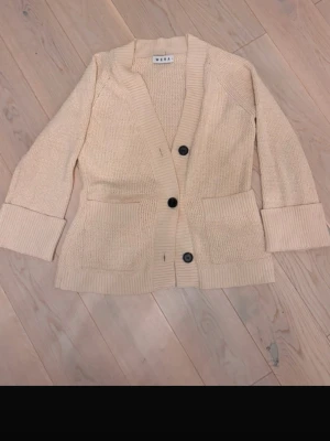 Beige stickad kofta från Wera - Beige stickad kofta från Wera med två stora fickor framtill och svarta knappar. Koftan har bred ribbad kant längs öppningen och ribbade muddar på ärmarna. Perfekt oversized känsla och mjukt material.