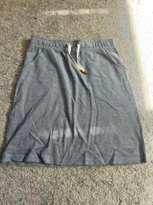 Grå kjol med snörning från Gant - Snygg grå kjol från Gant med vita snören i midjan och diskret logga broderad längst ner. Kjolen har en rak passform och är tillverkad i mjukt bomullsmaterial. Perfekt för en avslappnad och sportig stil.