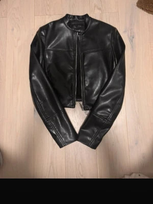 Svart skinnjacka från ZARA - Snygg svart skinnjacka från ZARA med rak passform och ståkrage. Jackan har dragkedja framtill och stilrena sömdetaljer över bröstet och ärmarna. Perfekt för dig som vill ha en klassisk och trendig look.