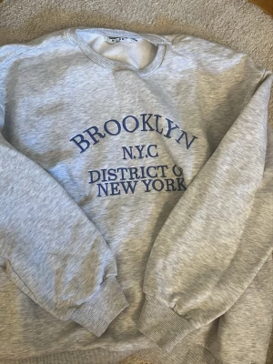 Collage tröja med tryck - Säljer en ljusgrå sweatshirt från WEEKDAY med blå text 'BROOKLYN N.Y.C DISTRICT OF NEW YORK' på bröstet. Tröjan har rund halsringning, långa ärmar och ribbade muddar. Perfekt för en chill och avslappnad stil.