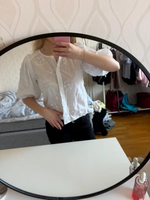 Vit blus med broderade blommor - Säljer en söt vit blus med broderade blommor och knäppning framtill. Blusen har rund halsringning och puffiga korta ärmar som ger en romantisk vibe. Perfekt att styla med jeans eller kjol för en trendig look.