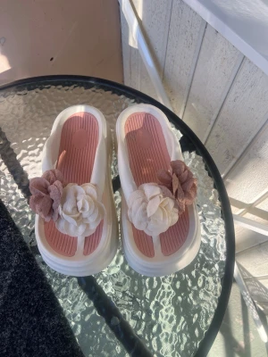 Vita sandaler med blommor - Säljer ett par chunky vita sandaler med rosa innersula och stora dekorativa blommor i vitt och ljusrosa framtill. Sandalerna har platåsula och är tillverkade i syntetmaterial. Perfekta för dig som vill sticka ut med en söt och unik detalj på fötterna.
