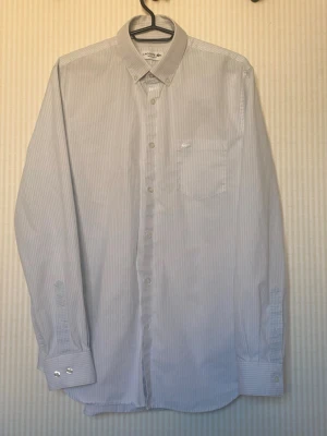 Vitblå randig skjorta från Lacoste - Klassisk skjorta från Lacoste i vitt och ljusblått med tunna ränder. Skjortan har lång ärm, knappslå framtill, bröstficka och button-down krage. Perfekt för en clean och stilren look.