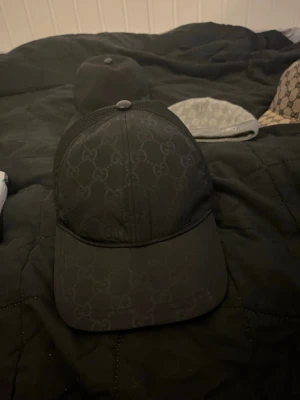 Svart keps från Gucci med GG-mönster - Säljer en svart keps från Gucci med det klassiska GG-monogrammet över hela framsidan. Kepsen har mesh på sidorna och baktill för extra ventilation samt justerbar rem bak. Perfekt för dig som vill ha en stilren och exklusiv accessoar.