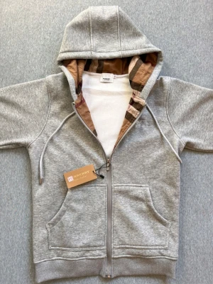 Grå hoodie från Burberry med dragkedja - Snygg grå hoodie från Burberry med klassiskt rutigt foder i huvan och silverfärgad dragkedja med Burberry-logga. Hoodien har snörning i huvan, två fickor fram och ribbade muddar. Perfekt för dig som vill ha en exklusiv och stilren look.