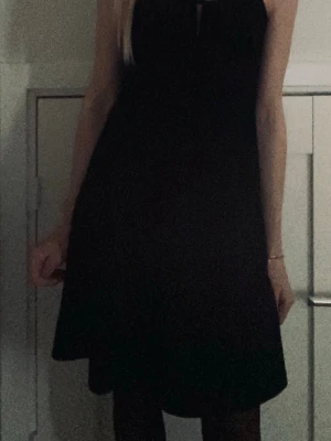 Svart kort klänning med smala band - Snygg svart kort klänning med smala axelband och en liten rosettdetalj framtill. Klänningen har en enkel, figurnära passform och är perfekt för dig som gillar minimalistisk stil. Materialet känns mjukt och följsamt.