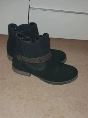 Svarta mockastövletter med rem - Säljer ett par svarta stövletter i mocka-look med dekorativ rem och knappdetalj på sidan. Skorna har låg klack och rundad tå, perfekta till jeans eller kjol. Klassisk och enkel design som passar till många olika stilar. Rieker 