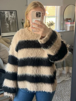 Beige och svart fluffig stickad tröja - Säljer en oversized stickad tröja med breda svarta och beige ränder. Tröjan är fluffig och har rund hals samt långa ärmar. Perfekt för dig som gillar en mysig och trendig look. Materialet känns mjukt och ger en avslappnad vibe.