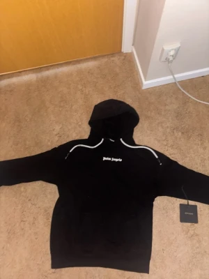 Svart hoodie från Palm Angels - Säljer en svart hoodie från Palm Angels med vit logga på bröstet och vita detaljer längs axlarna. Klassisk huva och känguruficka framtill. Perfekt för dig som gillar streetwear och vill ha en stilren men ändå unik look. Om ni hae npgra frågor skriv kan prata om priset privat. Finns defekter men inte stora.