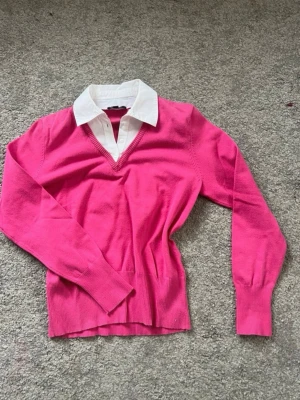 Rosa tröja med vit krage - Snygg rosa tröja med vit skjortkrage och v-ringning. Tröjan har långa ärmar och ribbade muddar vid ärmslut och nederkant. Perfekt för en preppy look och passar till både jeans och kjol.