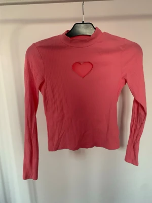 Rosa ribbad långärmad topp hjärtdetalj - Trendig rosa långärmad topp med ribbad struktur och ett hjärtformat utskärningshål på bröstet. Toppen har en högre hals och är figurnära i modellen, perfekt för dig som vill sticka ut med en söt detalj.