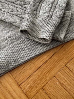 Grå kabelstickad tröja - Säljer en grå kabelstickad tröja med långa ärmar och ribbade muddar. Tröjan har ett klassiskt flätmönster längs ärmarna och är gjord i mjukt stickat material. Perfekt för dig som gillar en enkel men snygg look.