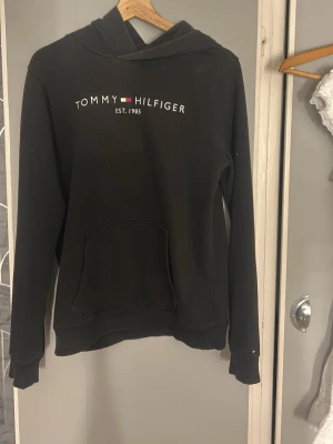 Svart hoodie Tommy Hilfiger - Svart hoodie från Tommy Hilfiger med logga och texten 'EST. 1985' broderat på bröstet. Klassisk känguruficka framtill, huva och ribbade muddar. Perfekt för en avslappnad och stilren look. Priset går att diskutera💕