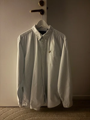 Randig skjorta från Lyle & Scott - Klassisk vit skjorta med tunna ljusblå ränder från Lyle & Scott. Skjortan har button-down krage, bröstficka med gul logga och knäppning framtill. Tillverkad i bomull och har lång ärm för en clean och stilren look.