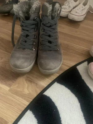 Gråa kängor med fuskpäls - Säljer ett par gråa kängor med snörning och mjuk fuskpäls på insidan. Kängorna har en robust sula och metallhål för snörena. Perfekta för kalla dagar när du vill hålla fötterna varma och snygga.