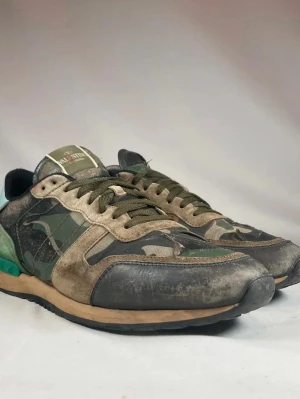 Valentino Rockrunners - Snygga sneakers från Valentino med camomönster i grönt, svart och beige. Skorna har detaljer i mocka och läder, samt en unik grön häl med nitar. Klassisk låg modell med snörning och svart sula.. De på undersidan som är smutsigt går att ta bort. Skorna är i st 44 men passar om man har st 45.