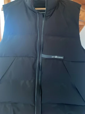 Svart dunväst från Sail Racing - Stilren svart dunväst från Sail Racing med hög krage och dragkedja framtill. Västen har två snedställda fickor och diskret logga på bröstet. Perfekt för lager-på-lager och håller dig varm under kyliga dagar.