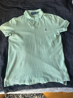 Ljusgrön pikéskjorta Polo Ralph Lauren - Snygg ljusgrön pikétröja från Polo Ralph Lauren med klassisk krage och två knappar framtill. Storlek står på bilden. Passar längdmässigt 170-178 typ. Tröjan har korta ärmar och broderade loggan på bröstet. Tillverkad i mjuk bomull som känns skön mot huden. Säljer då den är för liten, köptes ny på nk för 1 år sen. Varsamt använd och i väldigt fint skick.