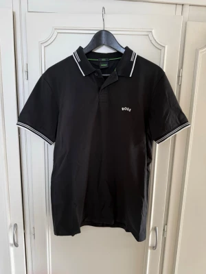 Svart pikétröja från BOSS - Klassisk svart pikétröja från BOSS med vit logga på bröstet och vita ränder på krage och ärmslut. Tröjan har korta ärmar, knappar vid halsen och är tillverkad i mjuk bomull. Perfekt för en clean och stilren look.