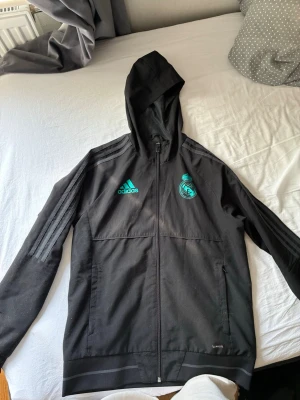 Svart Adidas Real Madrid vindjacka - Svart vindjacka från Adidas med huva och dragkedja. Jackan har Real Madrid-logga och Adidas-logga i turkos på bröstet samt klassiska ränder på axlarna. Tillverkad i polyester och har två sidofickor. Perfekt för dig som gillar sportig stil.