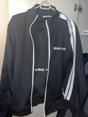 Palm Angels Tracksuit - Svart track jacket från Palm Angels med vita ränder längs ärmarna och logotyp på bröstet. Jackan har hög krage, dragkedja och ribbade muddar. Materialet är polyester och designen är sportig och modern. Superbra skick 9/10🌟 