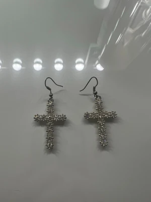 Korsörhängen med pärlor - Snygga örhängen i form av kors, dekorerade med små vita pärlor som ger ett glittrande intryck. Krokar i metall för enkel användning. Perfekta för dig som vill ha ett statement-smycke med unik design.