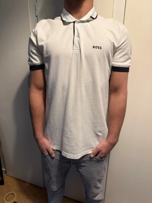 Vit pikétröja från BOSS - Vit pikétröja från BOSS med svart kant på ärmarna och diskret logga på bröstet. Klassisk krage med knappslå och en stilren passform som funkar till det mesta. Materialet känns mjukt och bekvämt. Nypris: 1000kr. Använd 2 gånger. Storlek: S men passar M.