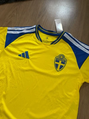 Sverige landslagströja - Säljer en gul svensk landslagströja från Adidas med blå detaljer och vita ränder på axlarna. Tröjan har broderad Sverige-logga på bröstet och Adidas-logga i blått. Tillverkad i lätt och ventilerande material, perfekt för fotboll eller träning. En snygg Sverigetröja nu inför VM2026.                                      Skriv om ni har några funderingar!😃
