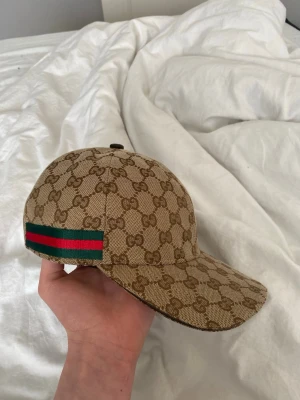 Gucci beige GG keps med röd/grön detalj - Säljer en beige keps från Gucci med klassiskt GG-monogram över hela kepsen. På sidan finns en ikonisk röd och grön rand. Kepsen har justerbart band bak och insidan är brun med Gucci-etikett. Materialet är canvas med bomullsfoder.