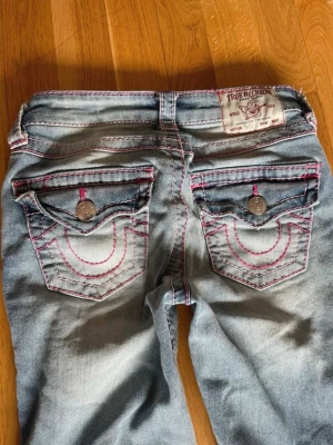 True Religion jeans - Säljer ett par ljusblå jeans från True Religion med ikoniska bakfickor och rosa kontrastsömmar. Jeansen har silverfärgade knappar på fickorna och en klassisk patch bak i midjan. Tillverkade i denim med snygg tvätt och detaljerad sömnad.