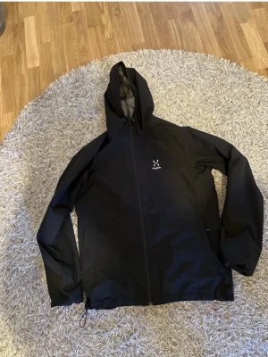 Svart vindjacka från Haglöfs - Snygg svart vindjacka från Haglöfs med minimalistisk design och hel dragkedja framtill. Jackan har huva och diskret logga på bröstet. Tillverkad i lätt och tåligt material som skyddar mot vind. Perfekt för dig som vill ha en stilren och funktionell jacka.