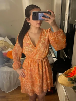 Orange omlottklänning med mönster - Säljer en kort omlottklänning i orange och vitt med abstrakt mönster. Klänningen har långärmade puffärmar, markerad midja med resår och v-ringad urringning. Tillverkad i ett lätt och luftigt material, perfekt för varma dagar.
