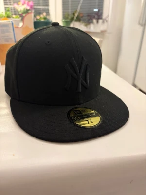 Svart New Era Yankees keps 59Fifty - Säljer en svart New Era 59Fifty keps med New York Yankees-logga framtill i svart brodyr. Klassisk flat brim och strukturerad passform. Kepsen är tillverkad i ett slitstarkt tyg och har den ikoniska New Era-loggan på sidan. Perfterk nu inför sommaren!