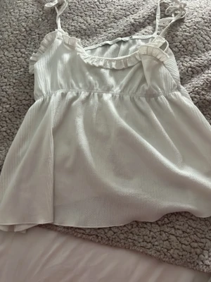 Vit linnetopp med volang från 157 - Söt vit linnetopp från 157 med tunna axelband och volangdetalj längs ringningen. Toppen har en lätt utsvängd passform och är tillverkad i ett mjukt, lätt material som passar perfekt till varma dagar. Enkel att styla med jeans eller kjol.