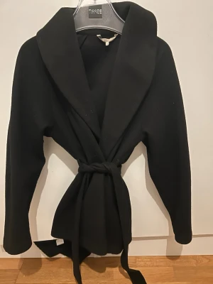 Svart kappa med bälte - Stilren svart kappa från obly med bred krage och knytskärp i midjan. Kappan har lång ärm och en klassisk, rak passform som ger en snygg siluett. 