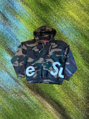 Camo hoodie från Supreme - Säljer en camouflagemönstrad hoodie från Supreme med dragkedja och stor vit Supreme-logga som går runt hela tröjan. Hoodien har huva med dragsko och ribbade muddar. Perfekt för dig som vill sticka ut med streetwear-vibe.