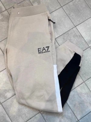 Emporio Armani EA7 byxor - Kvalitétsbyxor från Emporio Armani säljes  Snygga och bekväma byxor som passar både till vardags och helg. Ger en sportig, casual look. Slim men något stretch i materialet.  Klassisk EA7 logga på framsidan. Färgkombinationen är perfekt - beige, svart samt vit. Midjesnöret saknas.   Storlek M och passar längden 170-180cm  Nypris 1200kr