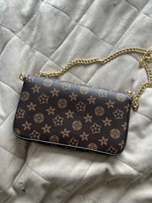 Brun vintage handväska med kedjerem - Snygg brun handväska med guldigt kedjehandtag och matchande plånbok. Väskan har ett klassiskt monogrammönster i beige och brunt, och är tillverkad i tåligt konstläder. Perfekt storlek för det viktigaste och enkel att bära över axeln.