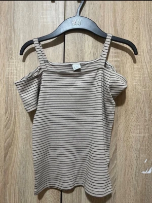 Beige randig offshoulder topp - Trendig beige topp med tunna svarta ränder. Offshoulder-modell med smala axelband och korta ärmar. Ribbad struktur som ger en snygg passform. Perfekt till jeans eller kjol för en vardagslook. Är aldrig använt passar 10-13 år 