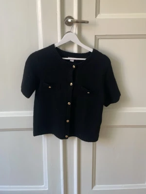 Svart stickad kortärmad kofta - Snygg svart stickad kofta med korta ärmar och rund hals. Koftan har guldfärgade knappar framtill och två små bröstfickor med dekorativa knappar. Perfekt att styla över en topp eller klänning för en trendig look.