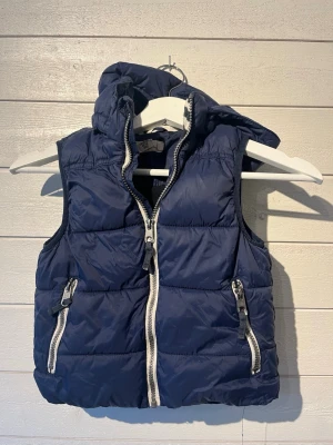 Mörkblå dunväst med huva Barn  - Snygg mörkblå dunväst med avtagbar huva och vita dragkedjor. Västen har två framfickor med dragkedja och quiltad design som ger en sportig look. Perfekt att slänga över en hoodie för extra värme under kyliga dagar.5-6 år . 