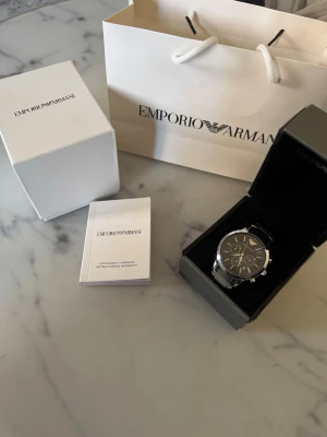 Emporio Armani klocka silver/svart - Stilren klocka från Emporio Armani med svart urtavla och silverfärgade detaljer. Klockan har kronograf, datumvisning och länkarmband i metall. Perfekt accessoar för dig som vill ha en klassisk och modern look. Kan göras mindre ifall du behöver det.