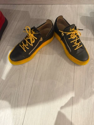 Svarta Giuseppe Zanotti sneakers med gula detaljer - Snygga svarta sneakers från Giuseppe Zanotti med gula skosnören och gul sula. Skorna har dragkedjor på sidorna och är tillverkade i läder för en exklusiv känsla. 