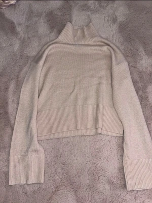Beige croppad stickad polotröja - Säljer en beige croppad stickad polotröja med bred ribbad kant nedtill och vid ärmslut. Tröjan har en hög krage och lång ärm, perfekt för lager på lager och en chill look. Passar till både jeans och kjol. Det finns bara en små fläck på handen som går säkert bort annars är tröjan i jättebra form, har inte använt den så många gånger