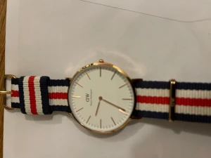 Daniel Wellington klocka randig NATO-band - Stilren klocka från Daniel Wellington med vitt ur och guldfärgad boett. Klockan har ett randigt NATO-band i marinblått, vitt och rött. Minimalistisk design med tunna index och DW-logga på urtavlan. Perfekt accessoar för en clean look.
