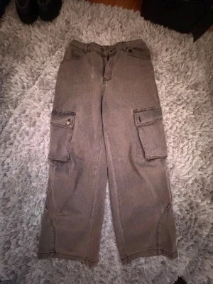 Grå cargobyxor med stora fickor - Säljer ett par grå cargobyxor med bred passform och stora fickor på sidorna. Byxorna har knapp- och dragkedjestängning samt detaljerade sömmar längs benen. Materialet är kraftigt och slitstarkt, perfekt för en avslappnad streetwear-look.