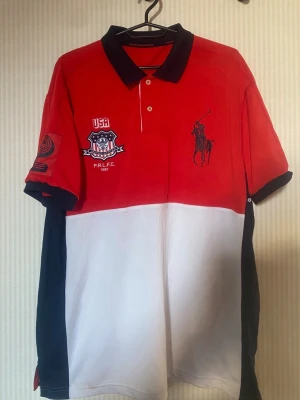 Röd och vit piké från Ralph Lauren - Säljer en snygg pikétröja från Ralph Lauren med röd ovandel, vit nederdel och svarta detaljer. Stora broderade loggor och USA-märke framtill, samt nummer 2 och USA-tryck på ryggen. Klassisk krage och knappar framtill. Perfekt för dig som gillar sportig stil.