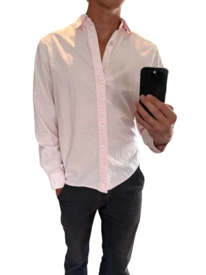 Ljusrosa skjorta med knappar från GANT - Säljer en stilren ljusrosa skjorta från GANT med klassisk krage och knäppning framtill. Skjortan har långa ärmar och är tillverkad i mjuk bomull som känns skön mot huden. Perfekt för dig som gillar en clean och enkel look.